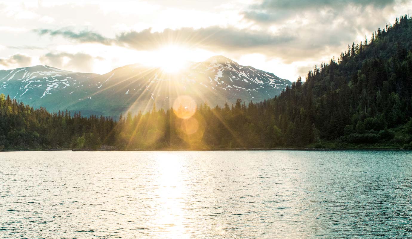 The sun shines from behind a lakeside mountain