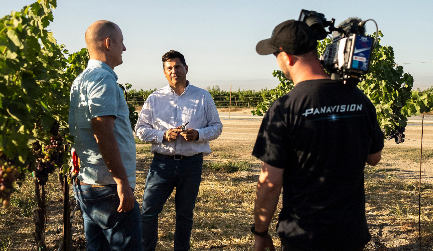 A cameraman stands with two men in a vineyard