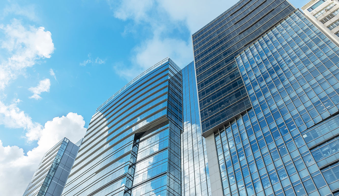 A modern cityscape featuring several tall glass-structured office buildings against a blue sky with scattered clouds.